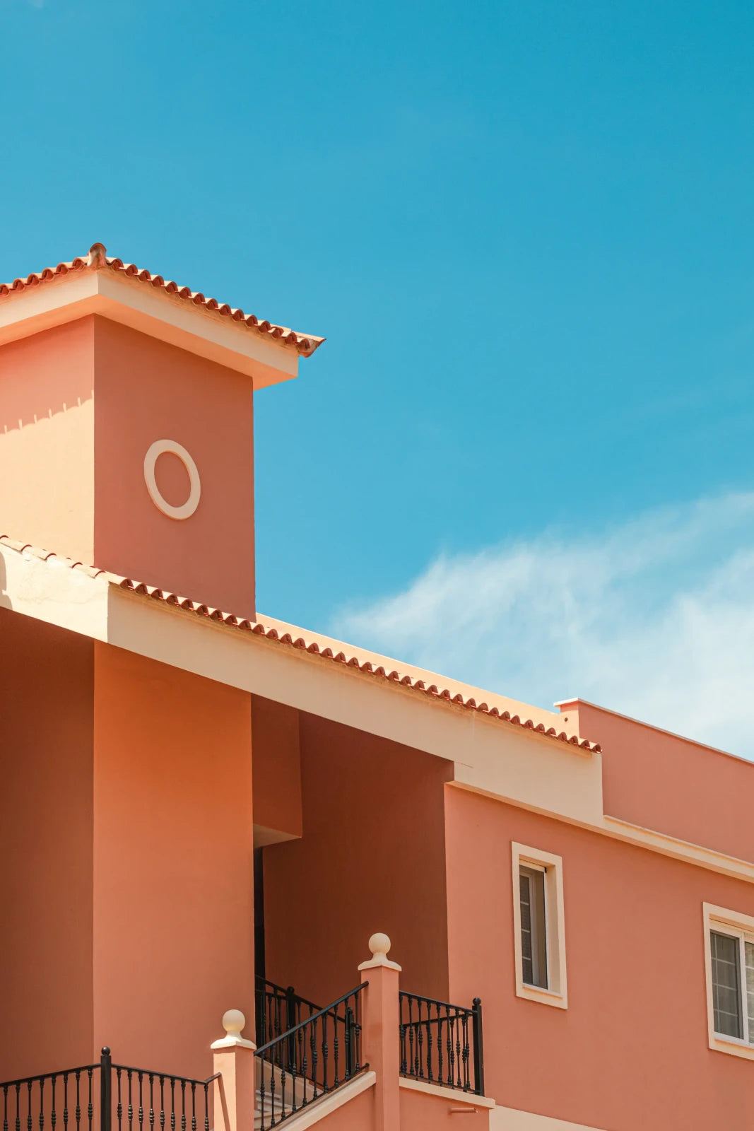 Coral-coloured building with pale roof trim, terracotta tiles and black railings against turquoise sky and a thin cloud.