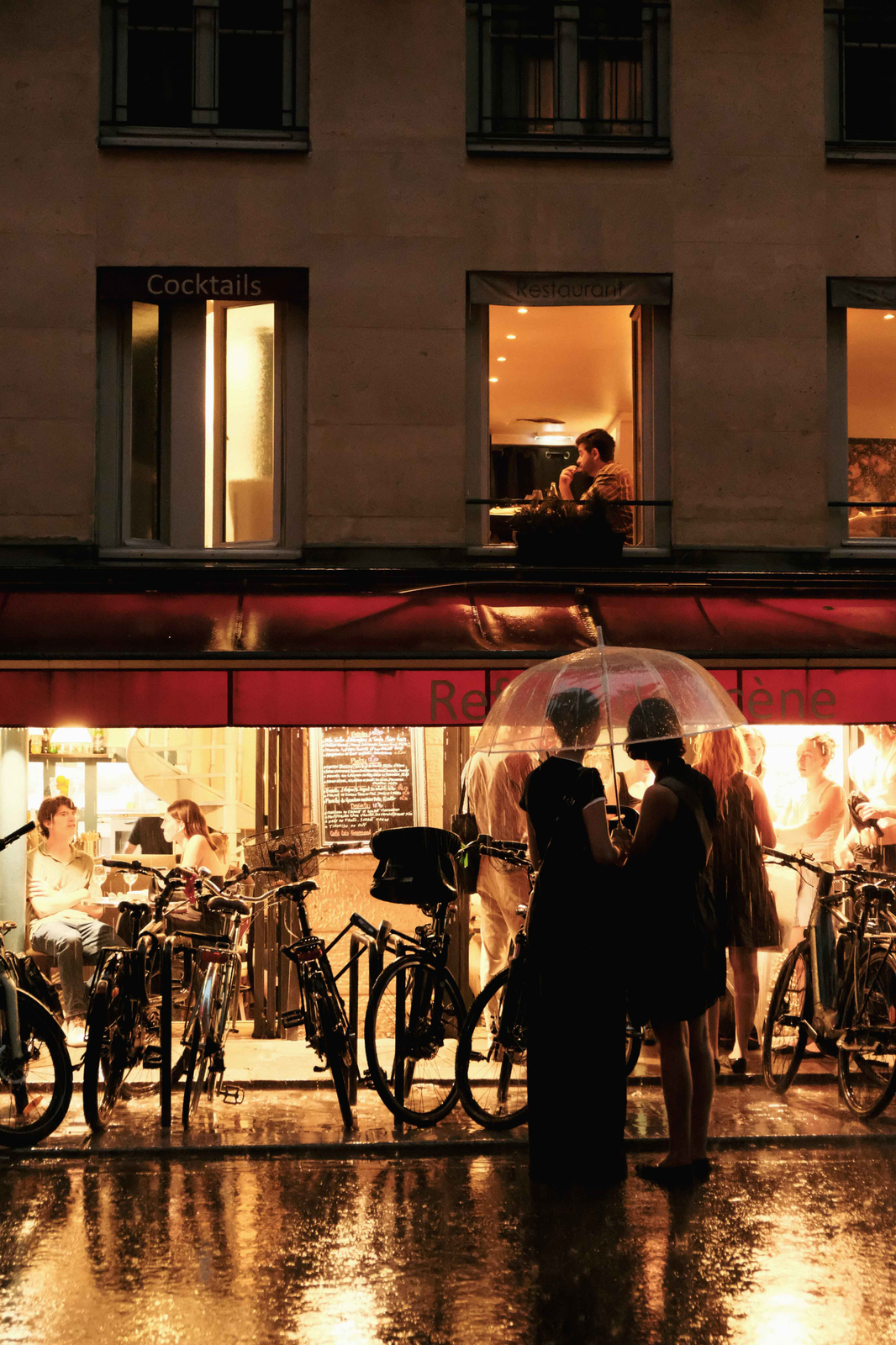 People shelter beneath a clear umbrella outside a brightly lit Paris café, with bicycles and warm reflections on the wet pavement.