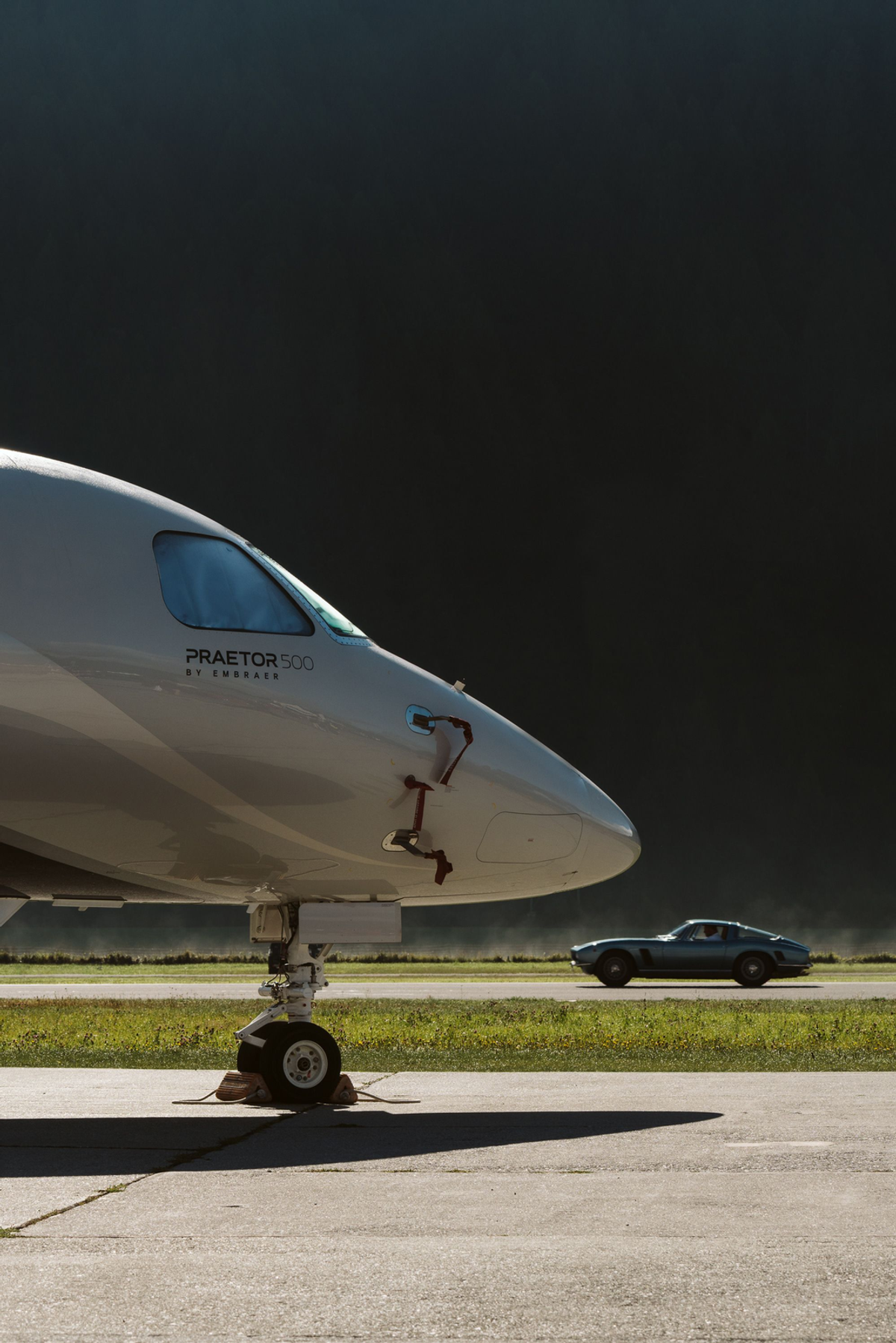 Close view of a silver private jet's nose, with a blue classic sports car behind it and a dark hillside beyond.