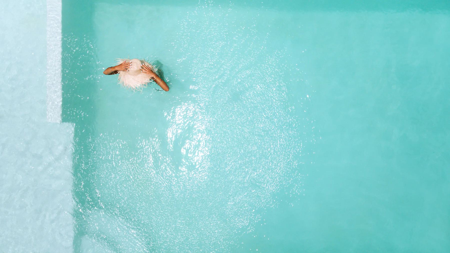 Top-down view of a person wearing a large, frayed straw hat, standing in bright, sunlit turquoise swimming pool water near white steps.