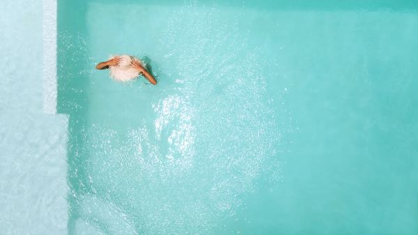 Top-down view of a person wearing a large, frayed straw hat, standing in bright, sunlit turquoise swimming pool water near white steps.