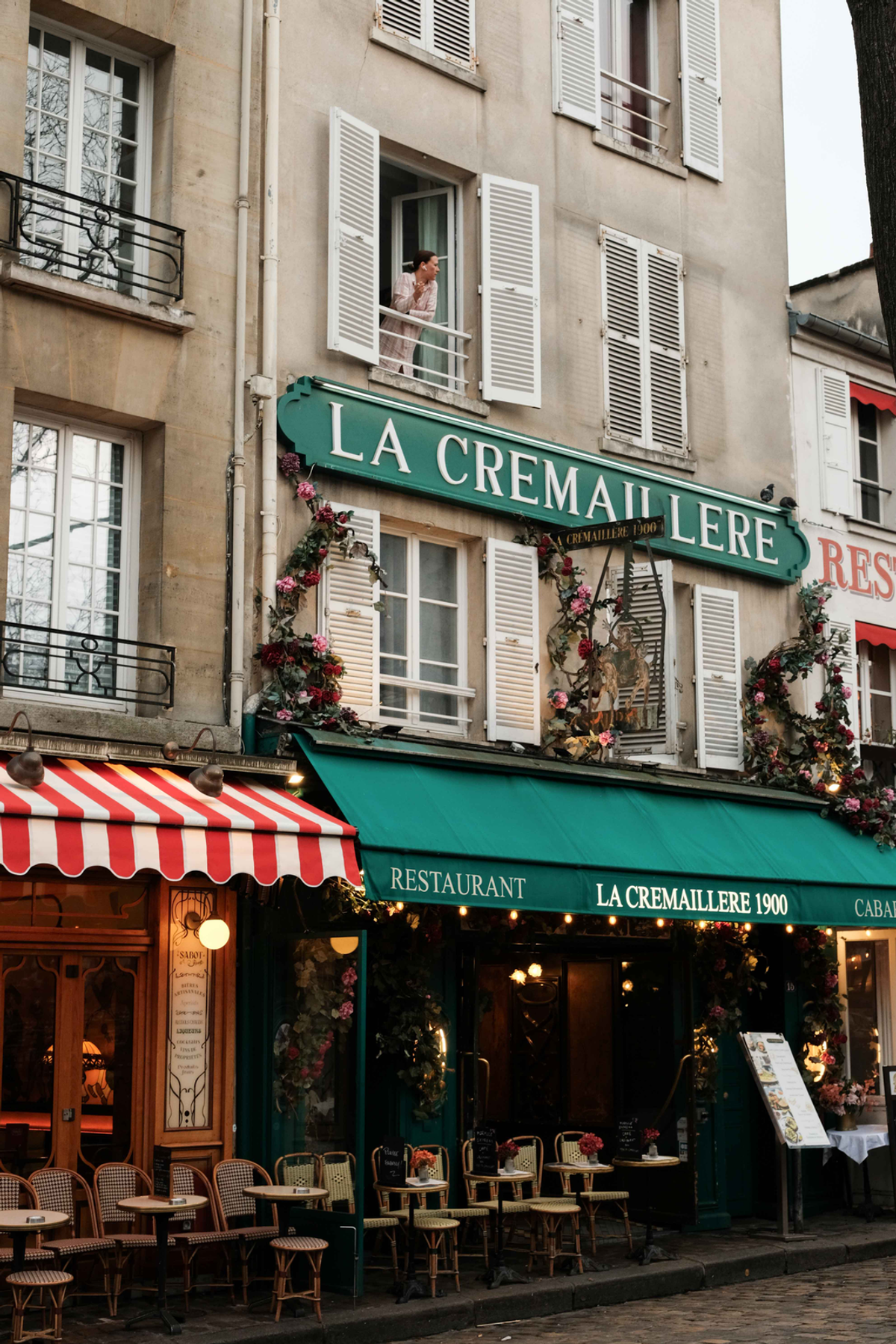A person leans from an open shuttered window above the green La Crémaillère restaurant sign, with café chairs and a red-and-white striped awning below.