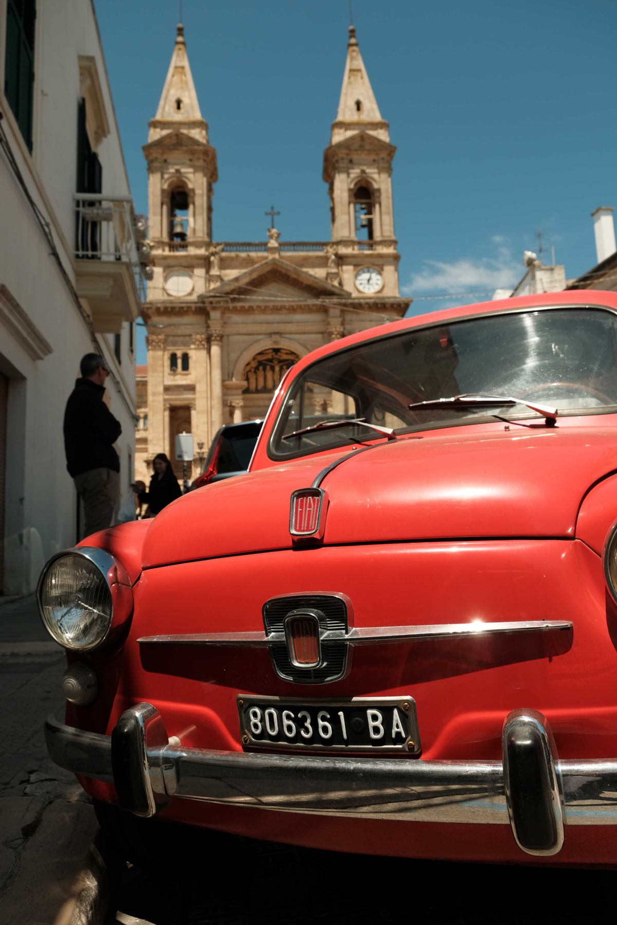 A bright red classic Fiat fills the foreground of an Italian street, with a stone church and two bell towers rising behind it under a blue sky.