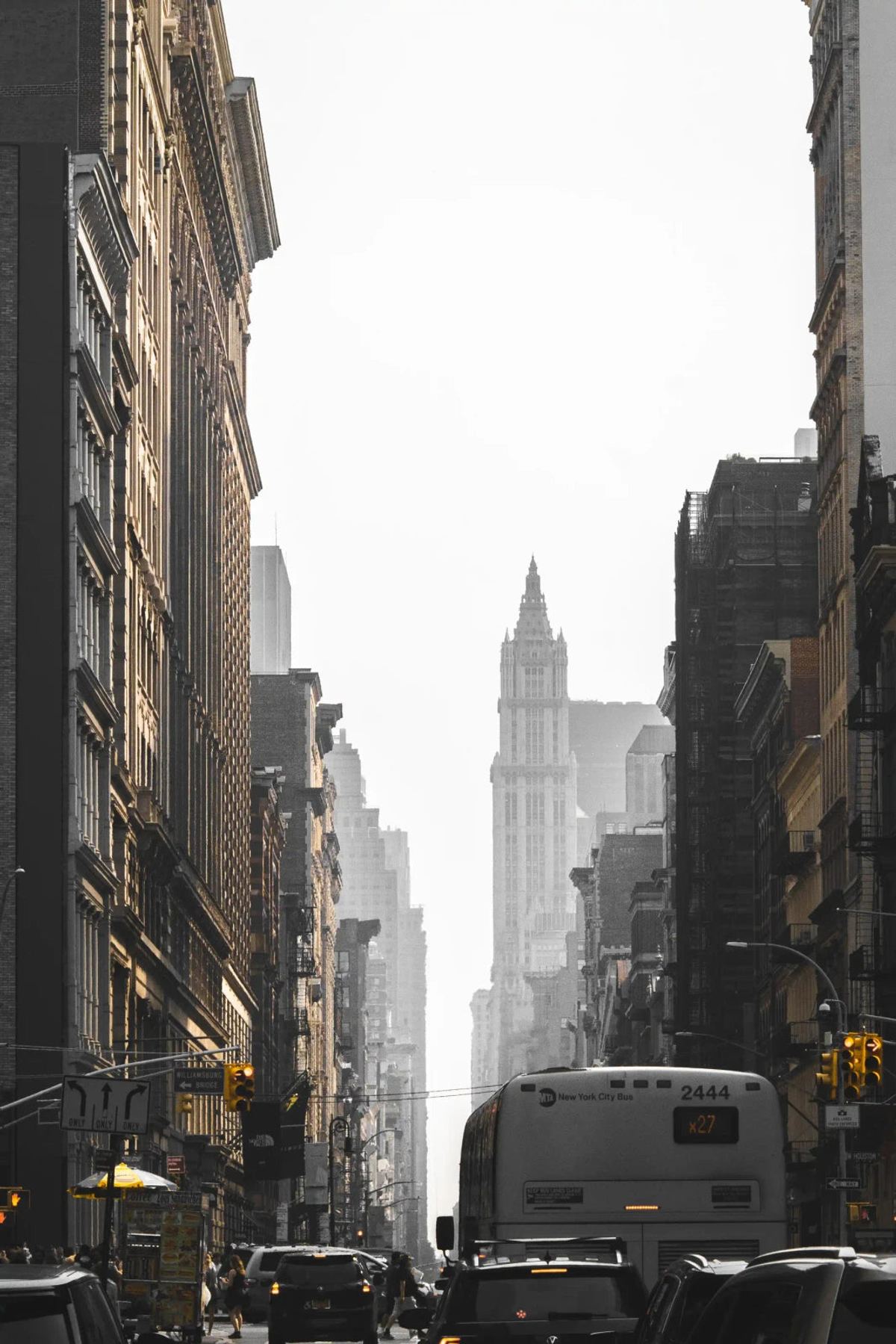 New York street with a bus, cars and yellow traffic lights between tall buildings, with a pale tower in the distance.