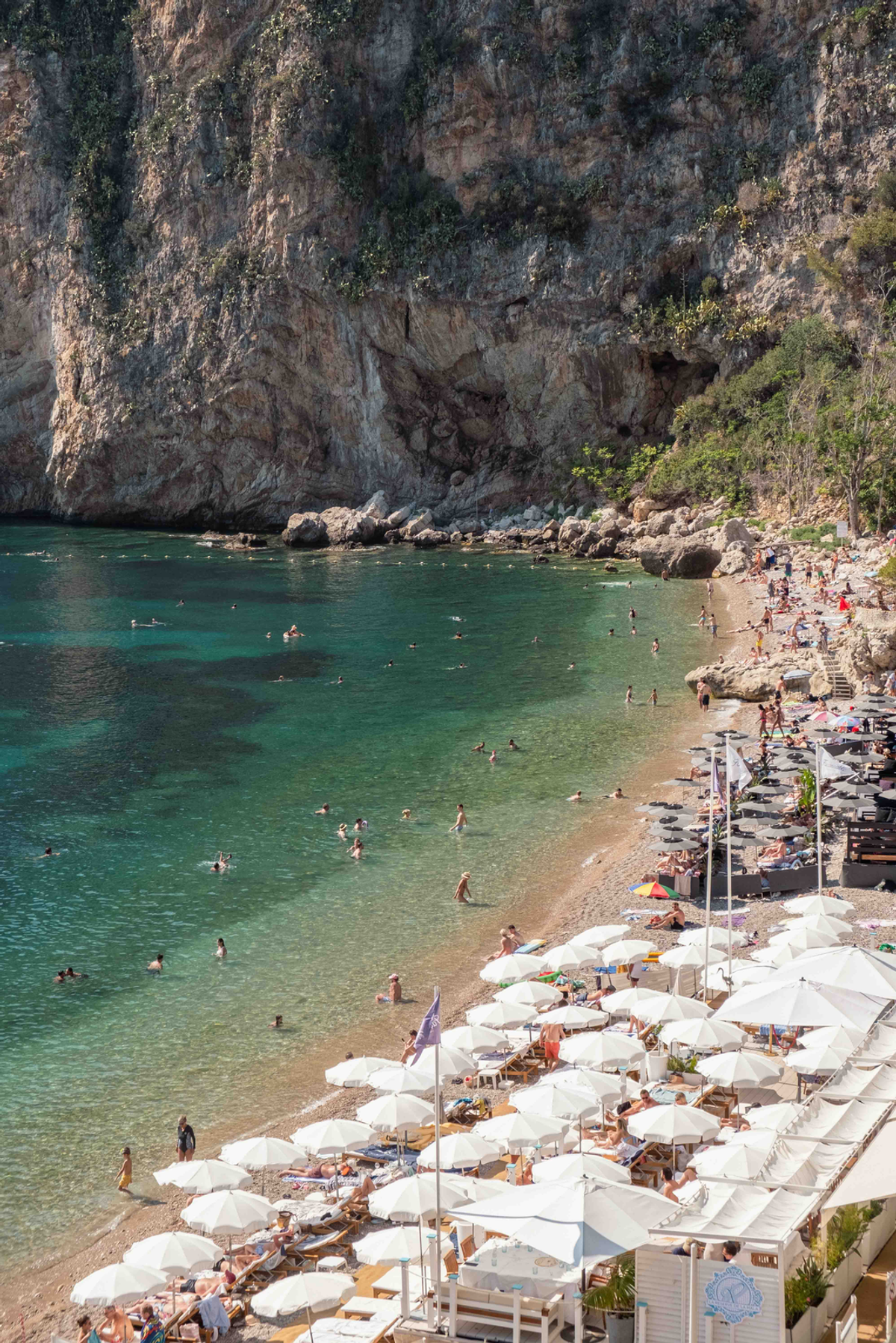 Rows of pale beach umbrellas follow the curve of a narrow Riviera beach beneath a cliff, with swimmers in the water.