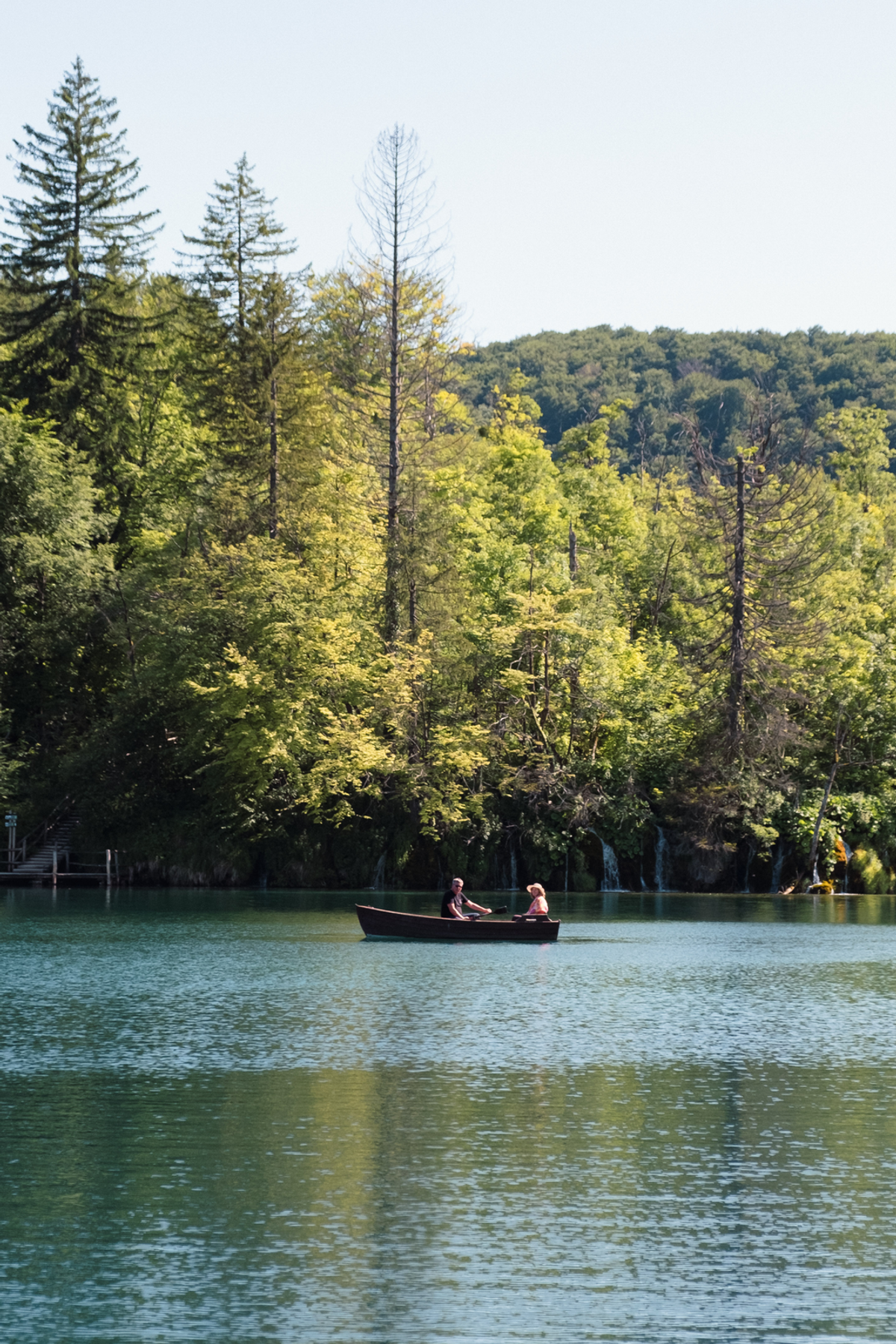 Two people in a small wooden rowing boat cross a green-blue lake beneath a wooded bank with slender waterfalls.