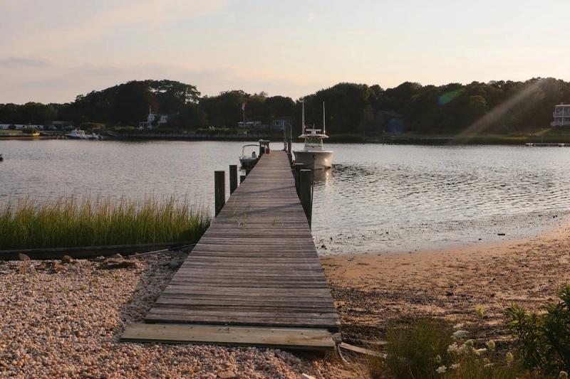 A long, weathered wooden jetty extending into a calm bay in the Hamptons