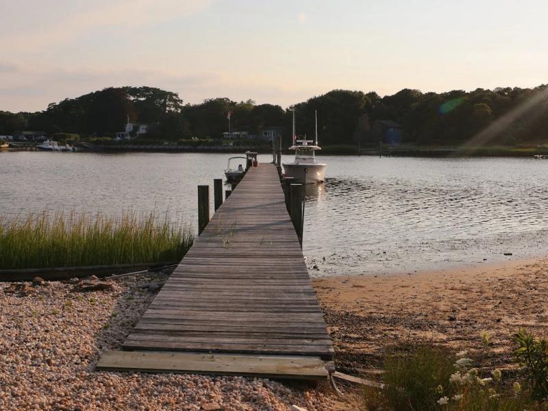 A long, weathered wooden jetty extending into a calm bay in the Hamptons