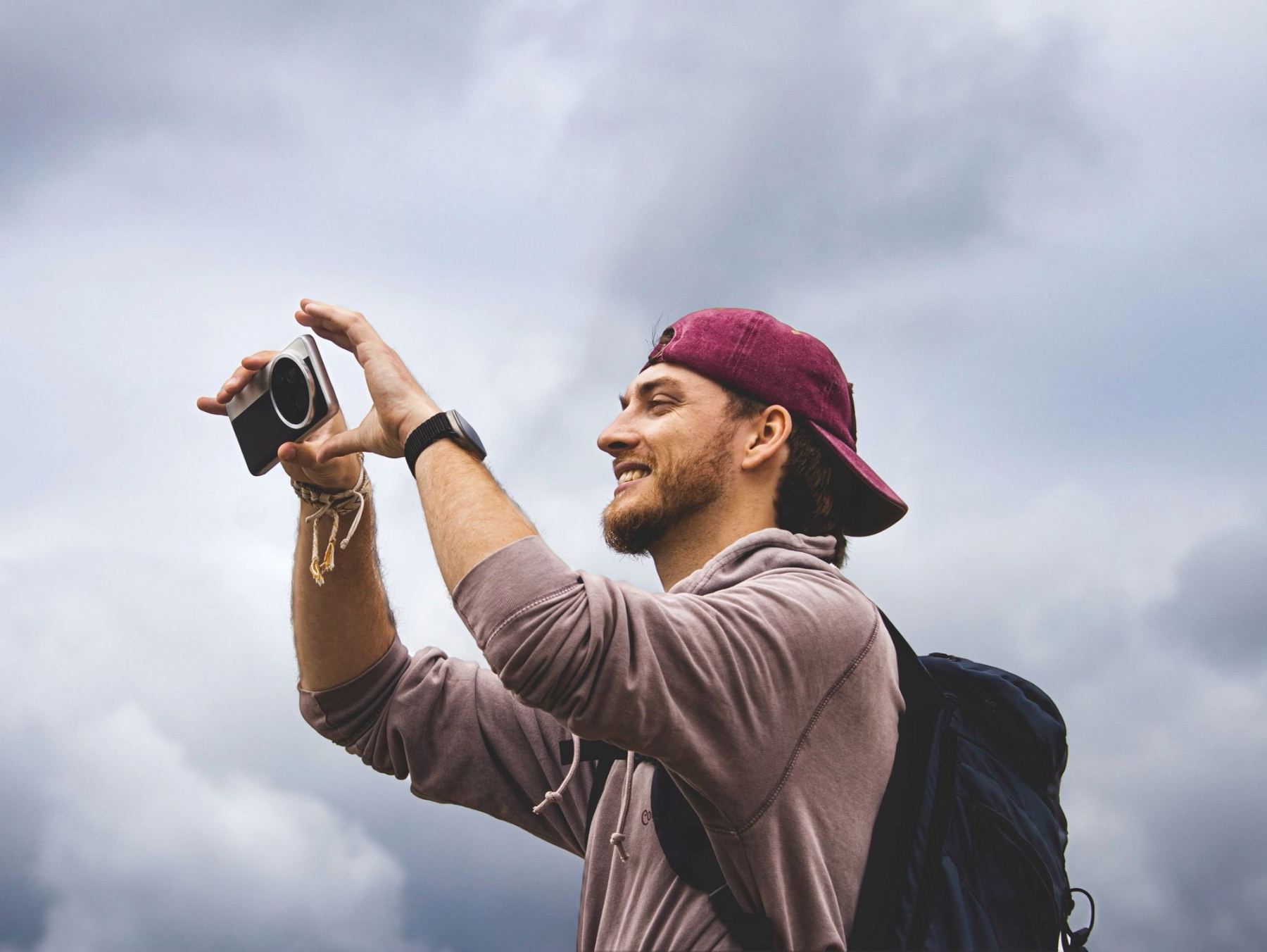 Man in a backwards red cap raising a compact camera against a dramatic overcast sky.