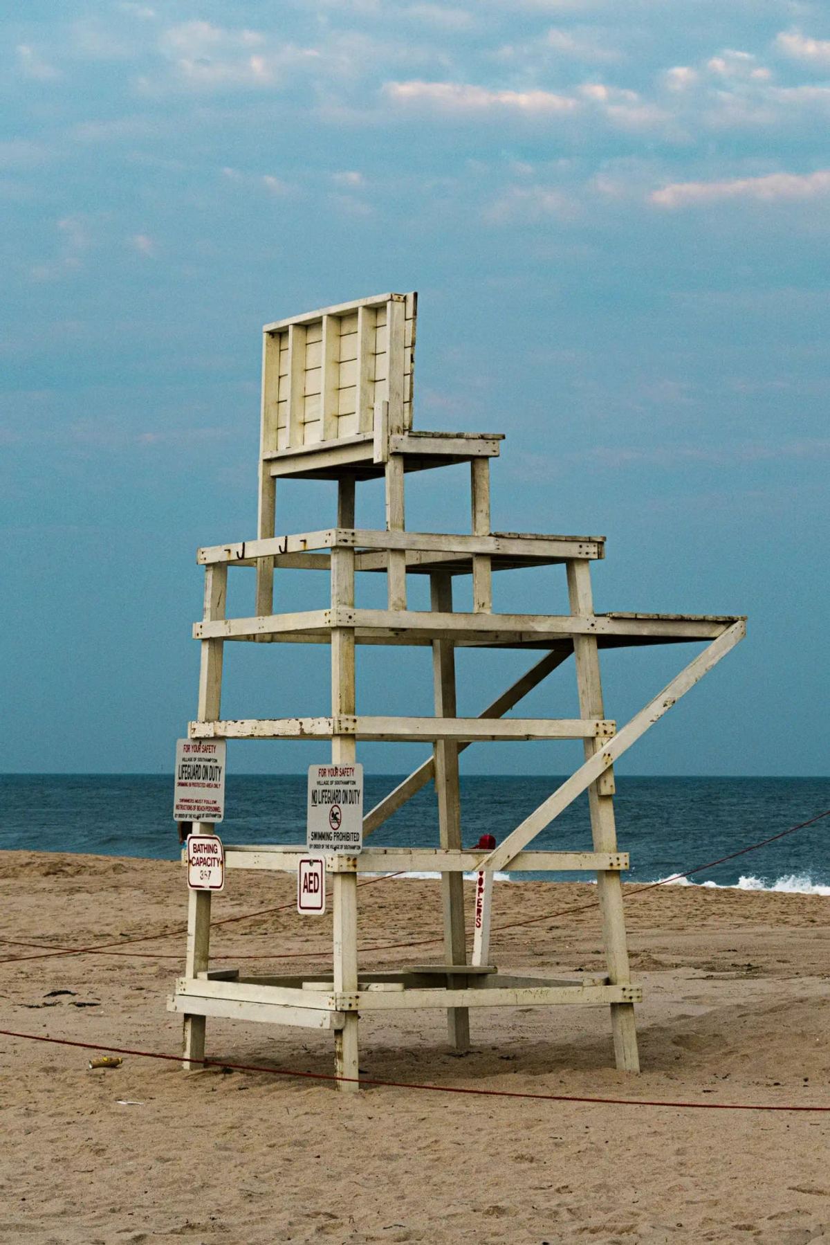 Empty, weathered wooden lifeguard chair on pale sand, with blue sea and lightly clouded sky behind it.