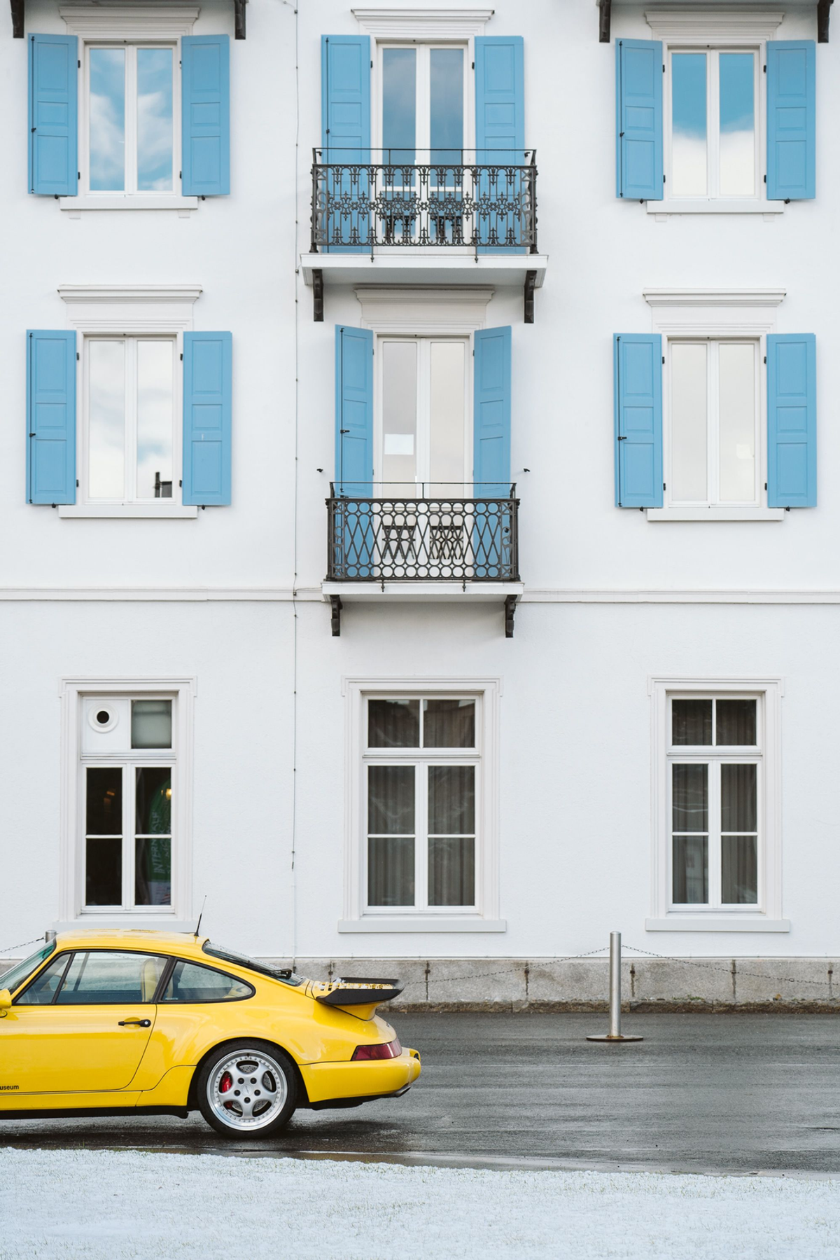 Yellow Porsche along a wet street beneath a white building with blue shutters and two ornate iron balconies, with snow in the foreground.