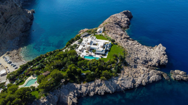 Aerial view of a white clifftop villa with a pool on a rocky peninsula above turquoise coves.
