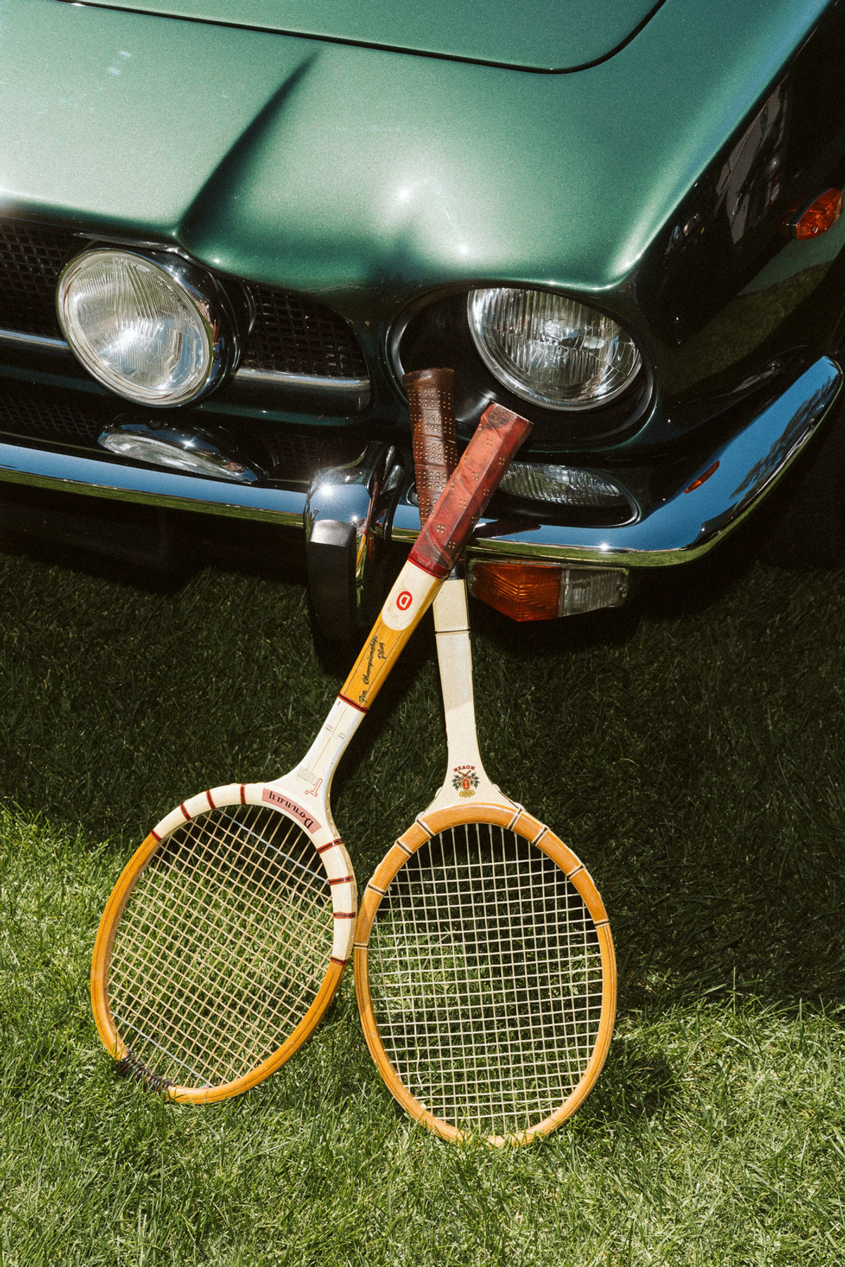 Two vintage wooden tennis rackets resting against the chrome bumper and round headlamps of a green classic car on grass.