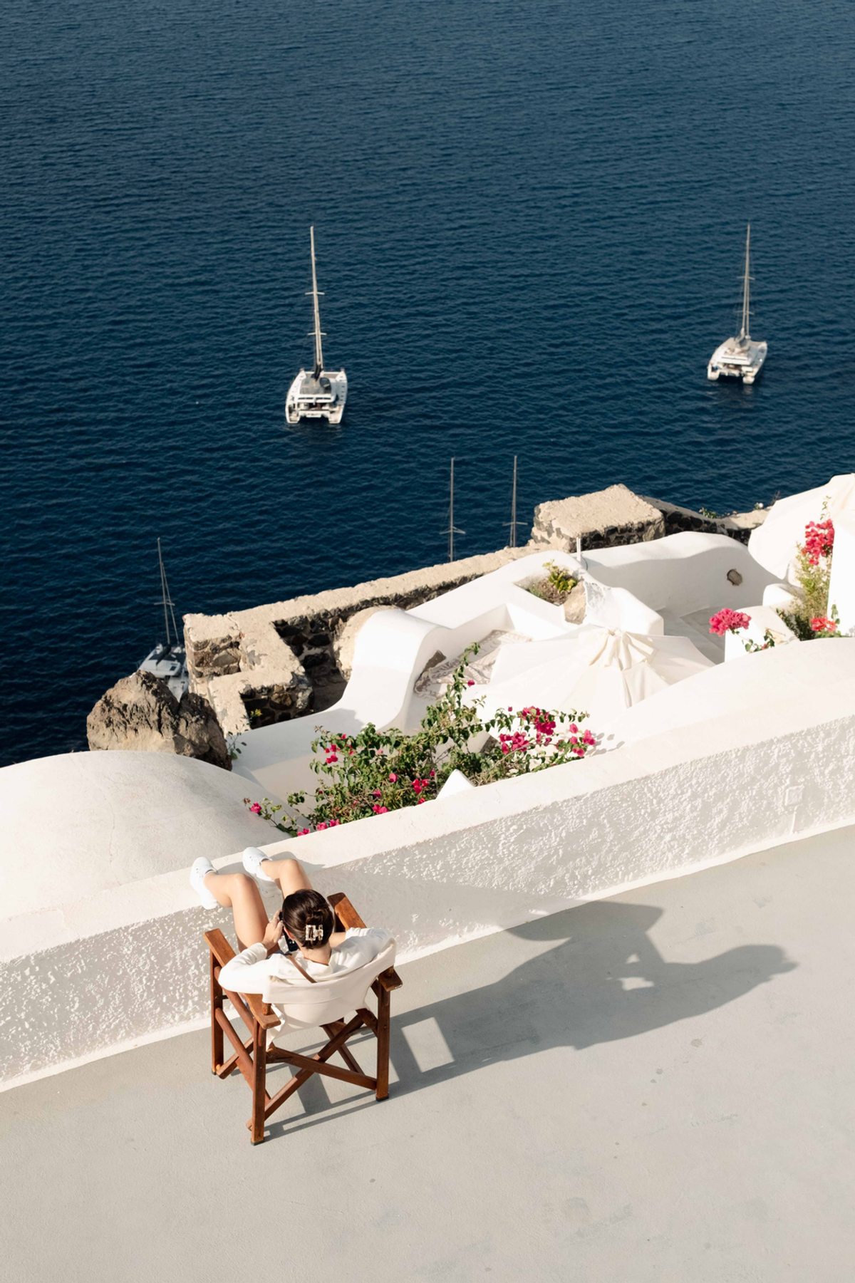Feet rest on a white terrace wall in Greece, with two boats offshore in deep blue water and pink flowers between the buildings.