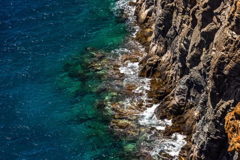 Clear blue-green sea and white foam meet an angular, fractured rock face along a diagonal shoreline.