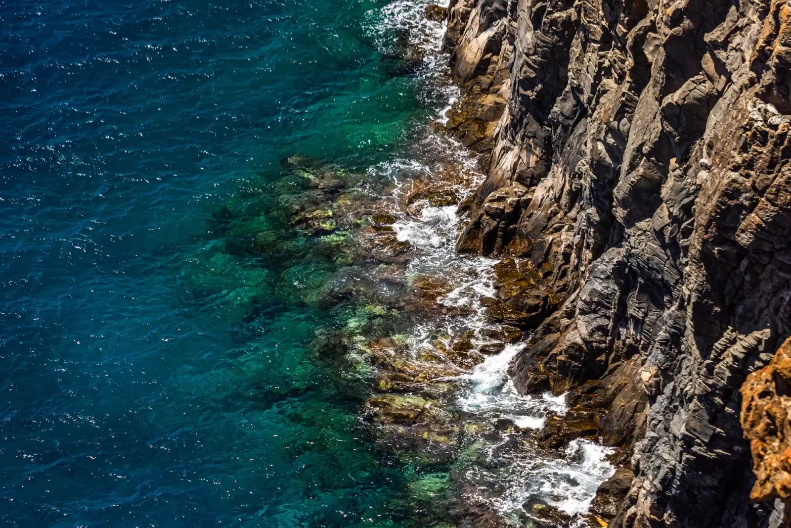 Clear blue water meets a steep, fractured rock face, seen from above, the shoreline cutting diagonally across the frame.
