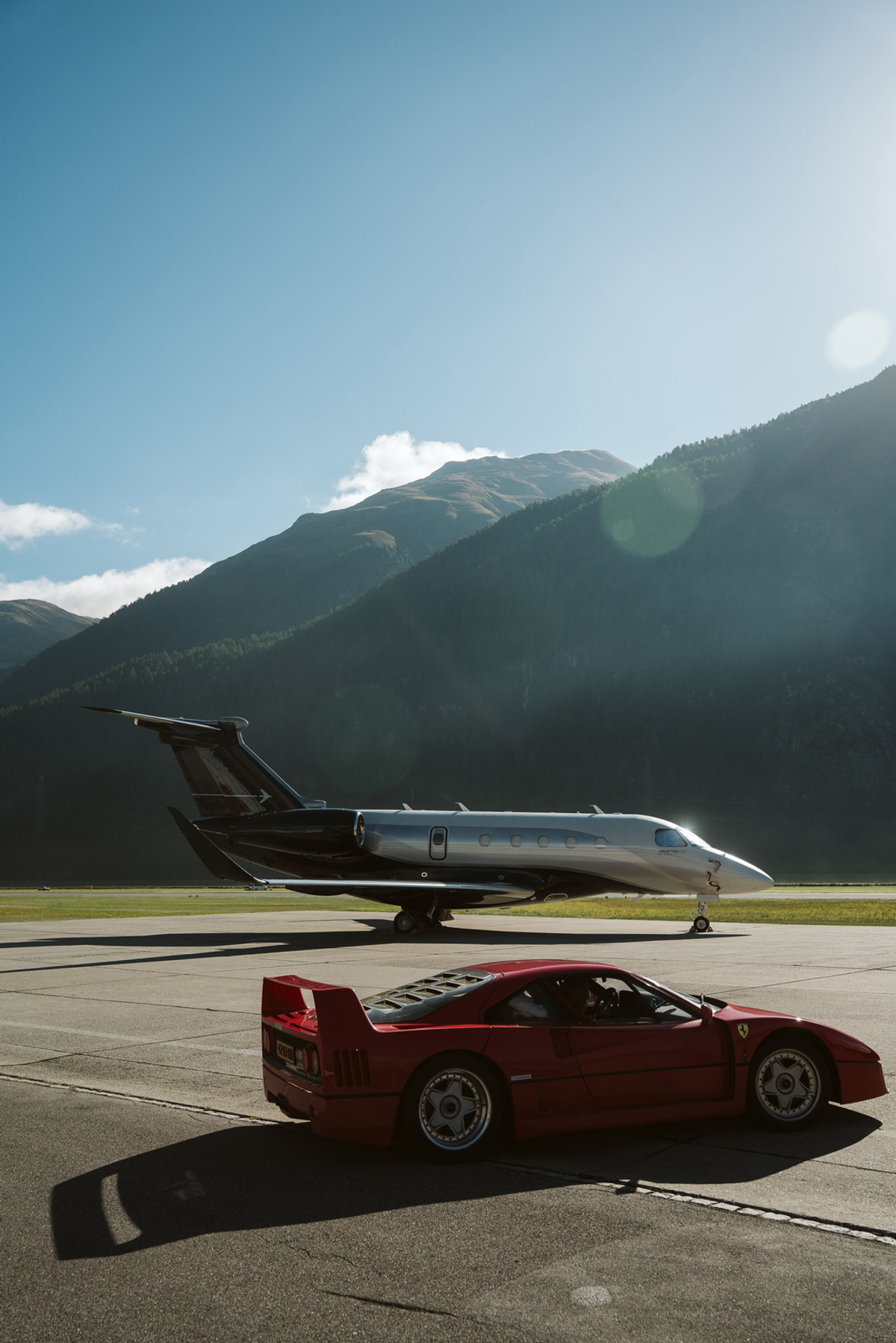 Red Ferrari F40 and silver private jet on an airfield beneath forested mountains and a clear blue sky.