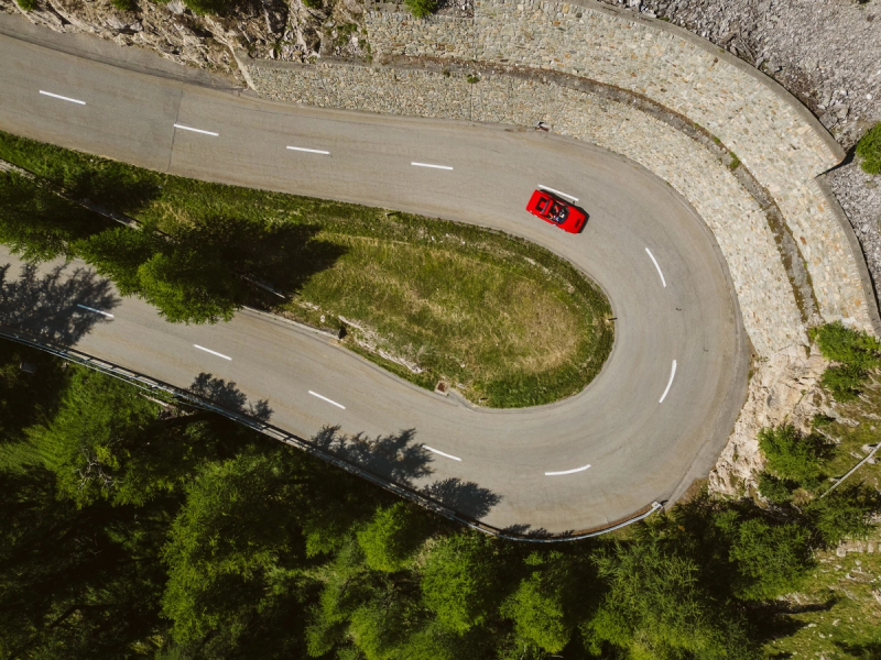 Aerial view of a red convertible rounding a mountain hairpin bordered by stone walls, grass and green trees.