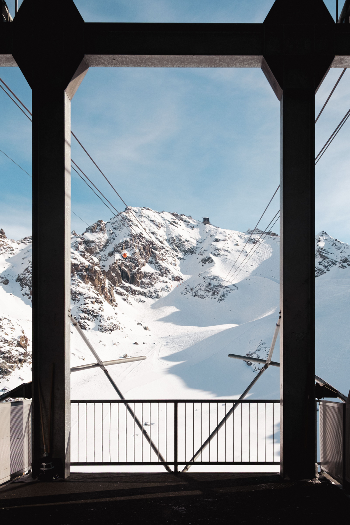 Dark structural beams frame a snowy mountain view from a cable-car station, with cables leading towards the peaks.