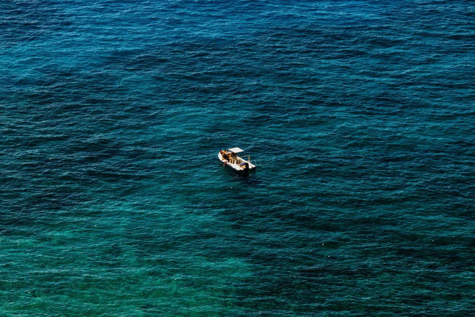 A small boat with a white canopy surrounded by rippling turquoise, teal and deep blue water, seen from above.
