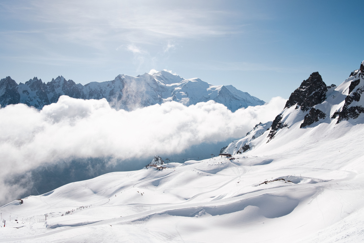 Sunlit snowfields and dark mountain peaks sit above a low bank of cloud, with distant ridges beneath a blue sky.