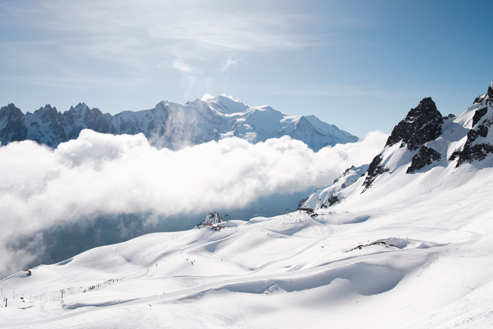 Sunlit snowfields and dark mountain peaks sit above a low bank of cloud, with distant ridges beneath a blue sky.