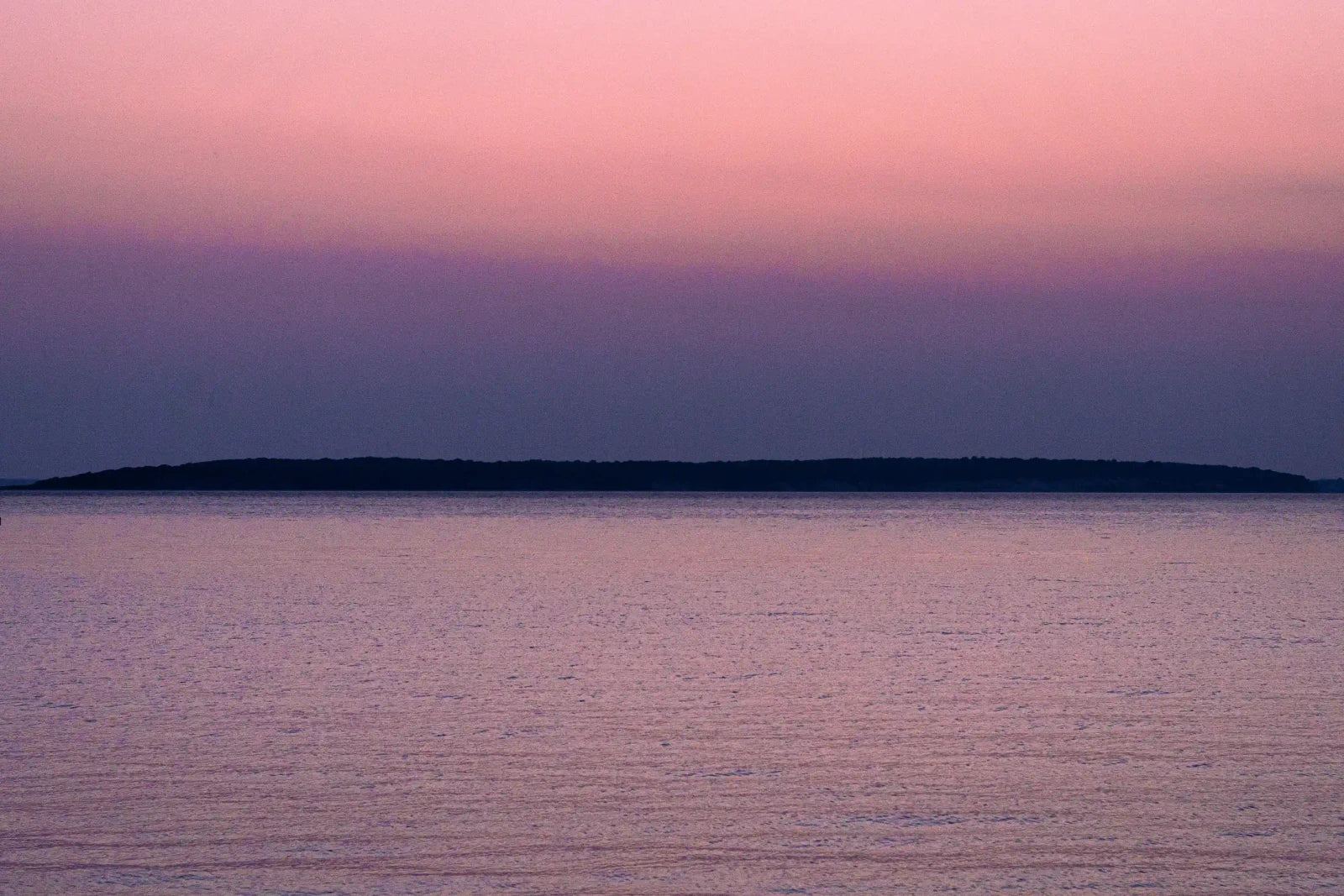 Pink light settles across open water, with a low strip of land holding the horizon.