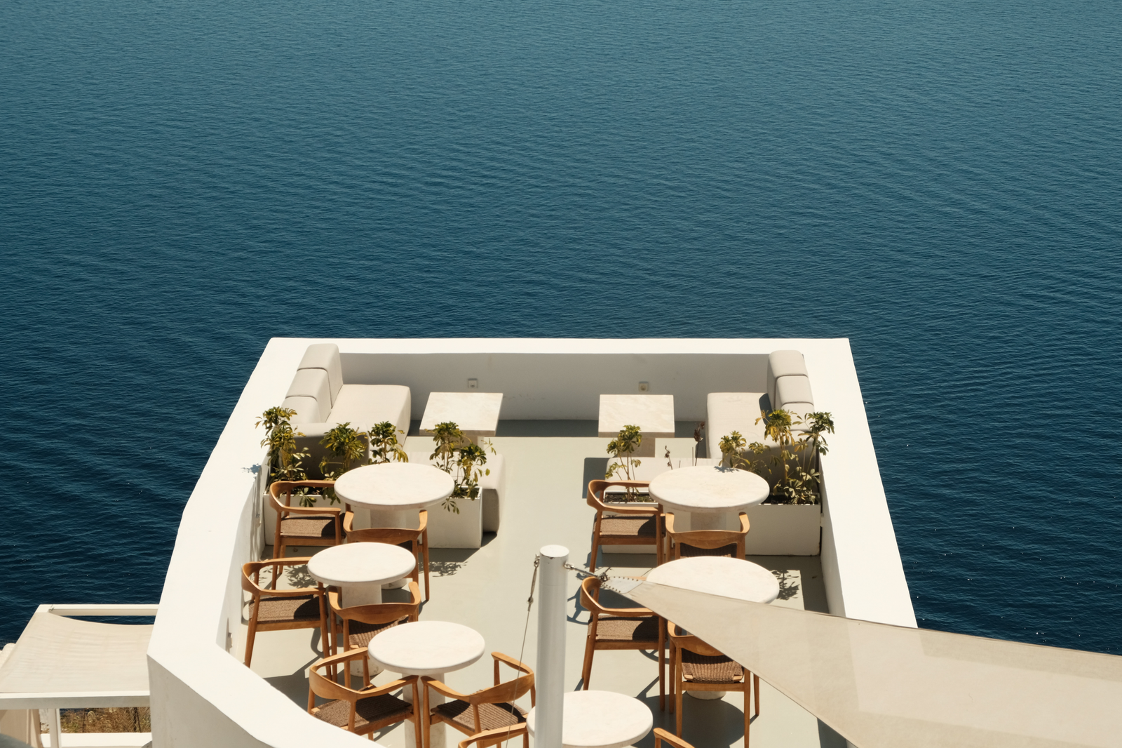 Round tables and curved wooden chairs on a white terrace that projects into an uninterrupted field of blue sea.