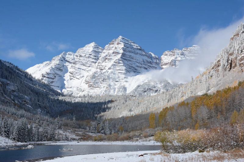 Snow-capped Maroon Bells peaks above a calm lake with autumn golden aspens and evergreen forest