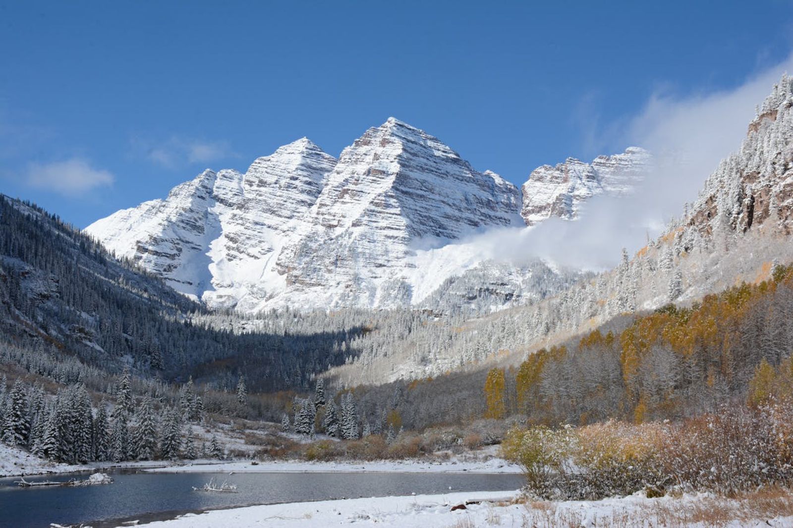 Snow-capped Maroon Bells peaks above a calm lake with autumn golden aspens and evergreen forest