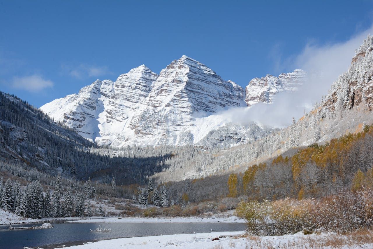 Snow-capped Maroon Bells peaks above a calm lake with autumn golden aspens and evergreen forest