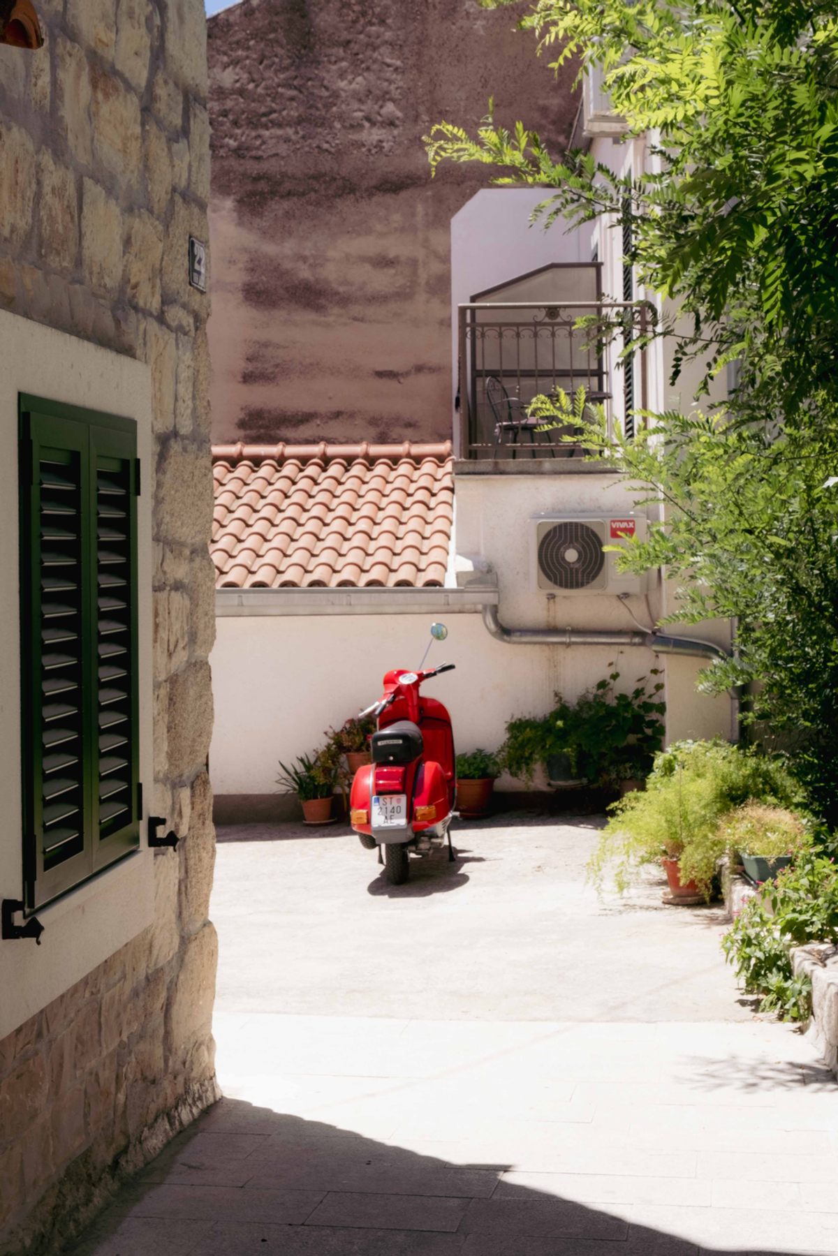 A red scooter stands at the end of a stone passage in Croatia, framed by shutters, roof tiles and potted plants.