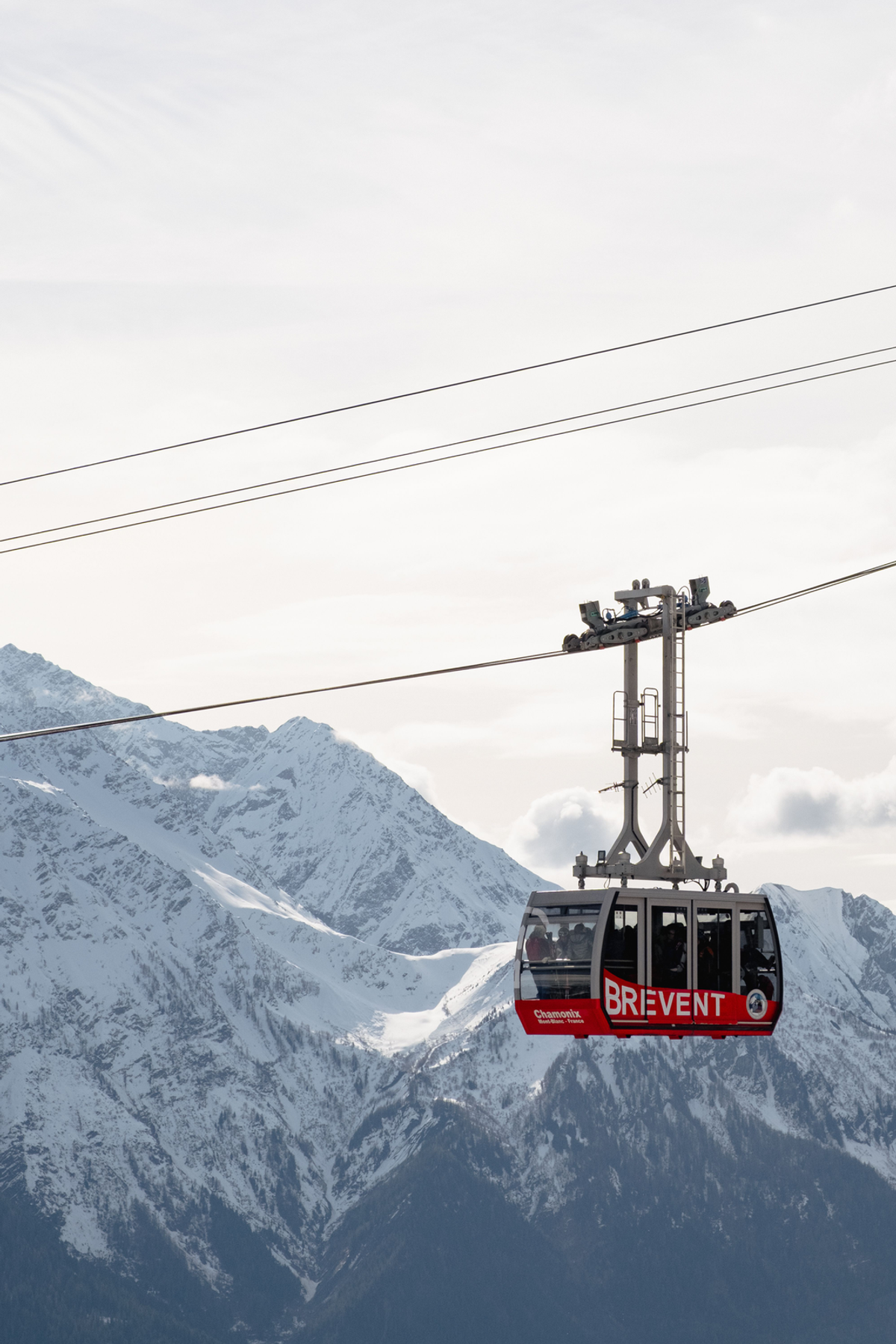 A red cable car travels along diagonal cables in front of snowy mountain peaks and a pale winter sky.