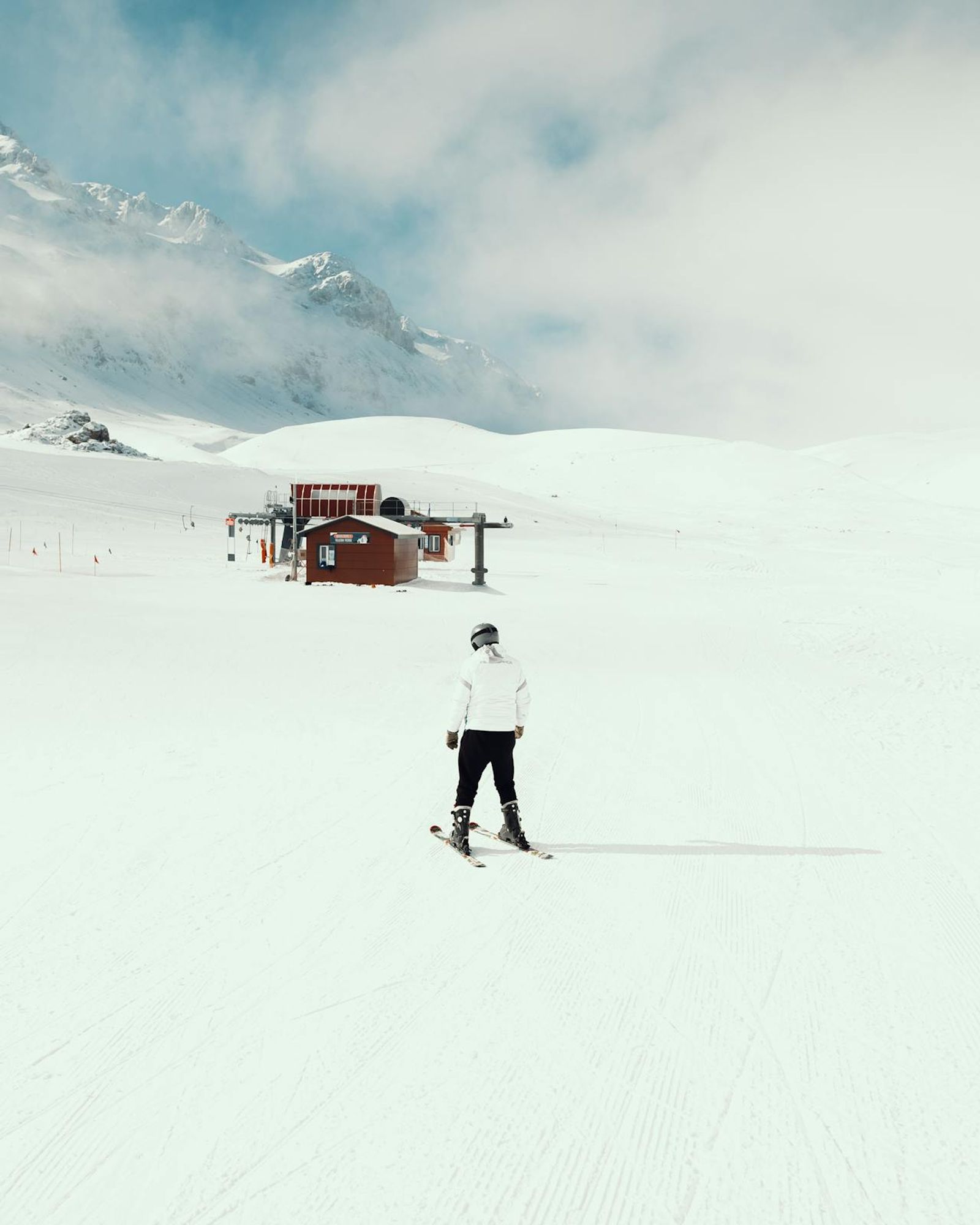 Skier in white jacket on a groomed piste approaching a red ski lift station under a cloudy peak