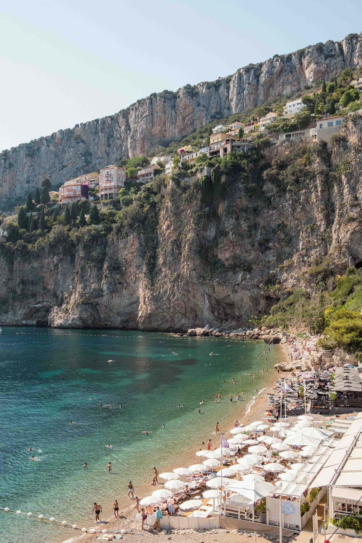 White parasols gather along the curve of a Côte d’Azur beach beneath great faces of rock, with swimmers in clear green water.