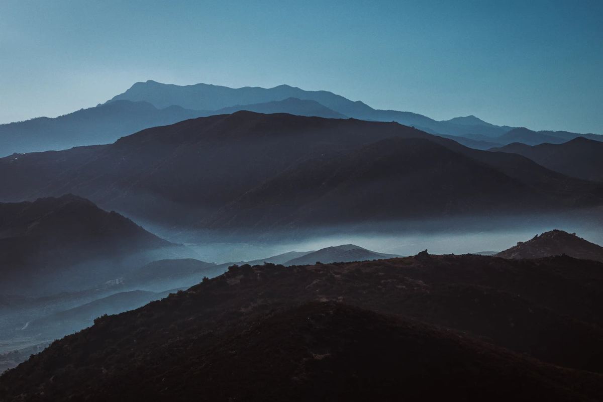 A dark ridge crosses the foreground beneath successive blue mountain silhouettes and narrow bands of valley mist.