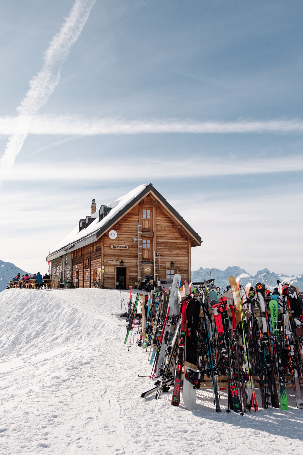 A timber mountain hut stands in bright snow beneath a blue sky, with a crowded rack of skis in the foreground.