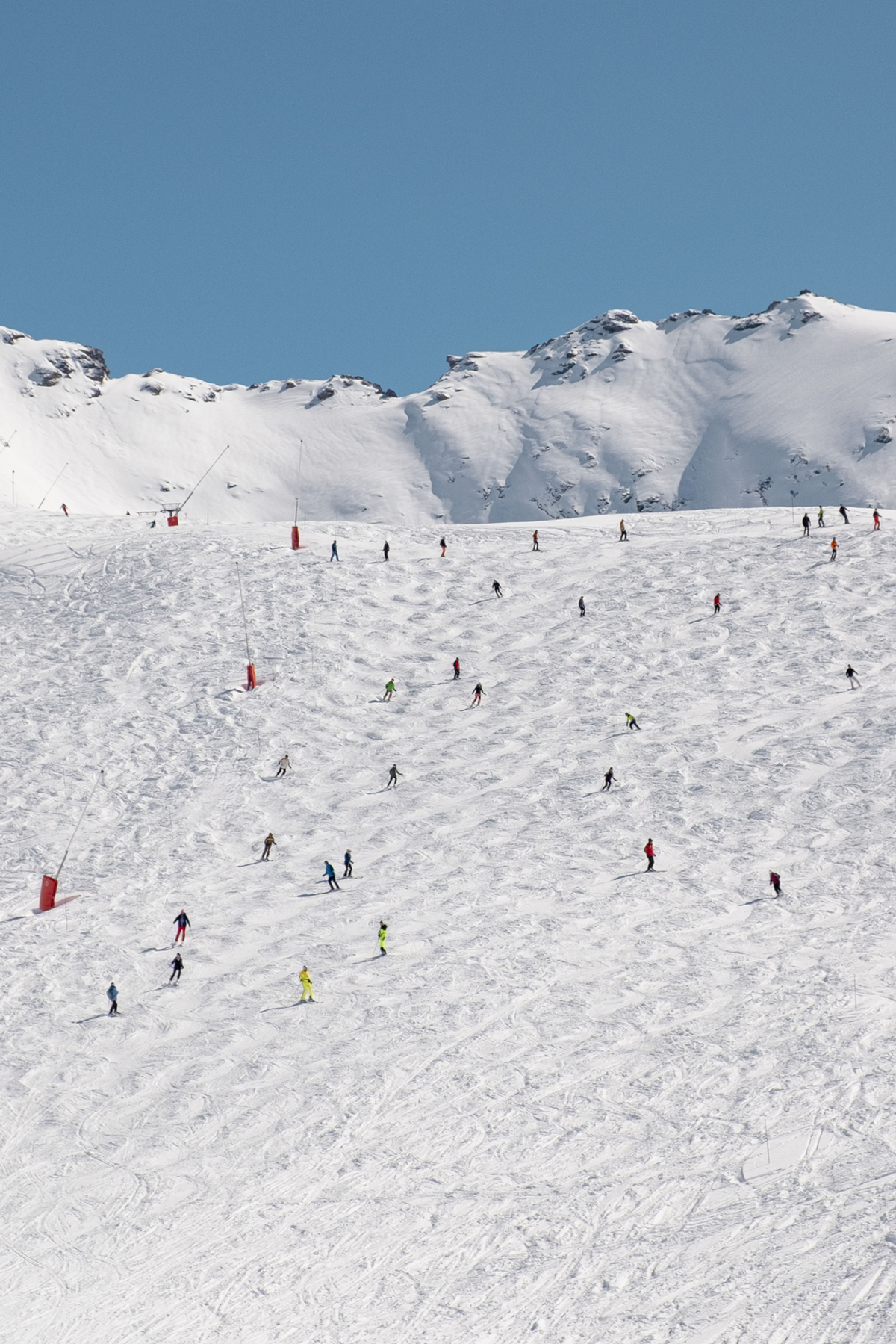 Many small skiers scatter across a broad sunlit piste below a snowy ridge and a clear blue sky.