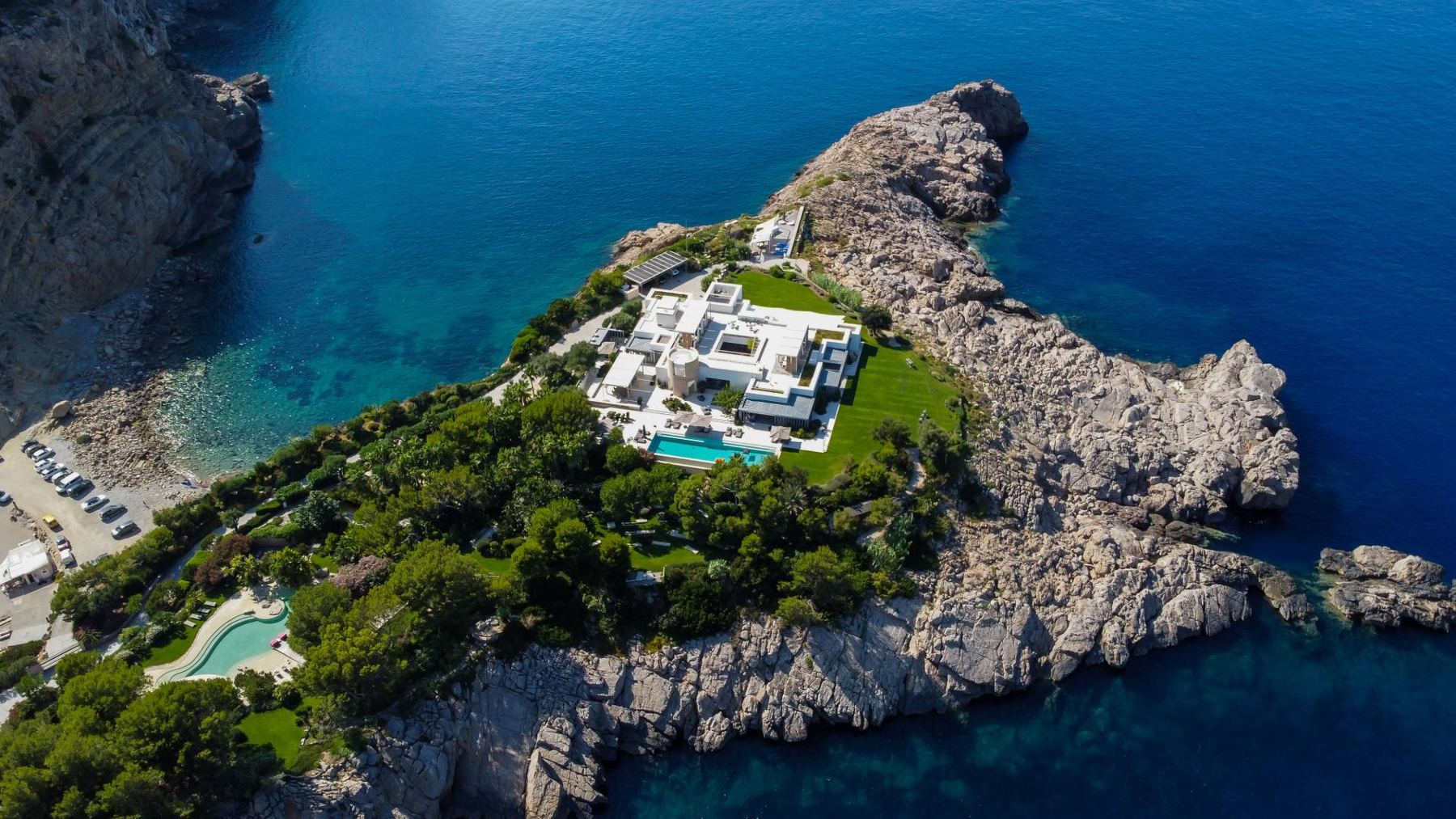 Aerial view of a white clifftop villa with a pool on a rocky peninsula above turquoise coves.