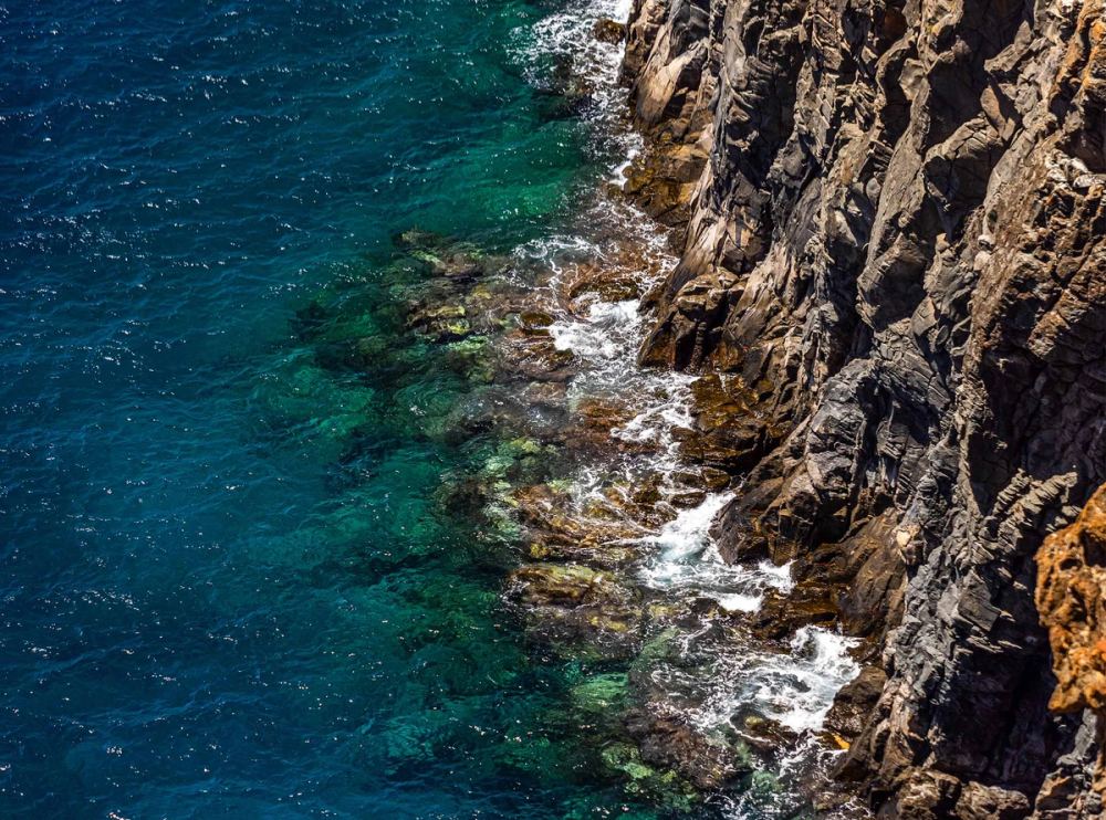 Rugged cliff face meeting clear turquoise water, seen from above.