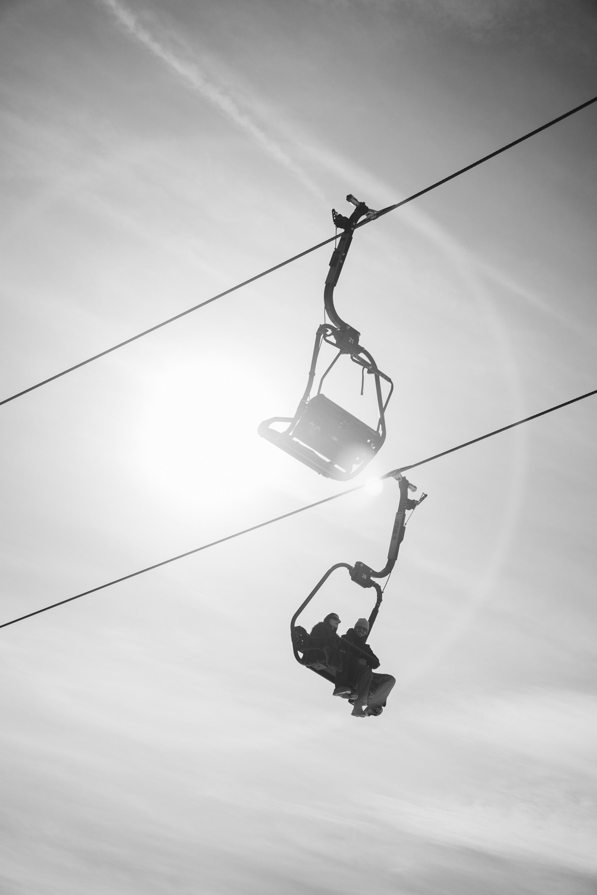 Black-and-white photograph of an empty chairlift and a second chair carrying two people, silhouetted against bright sun and a pale sky.
