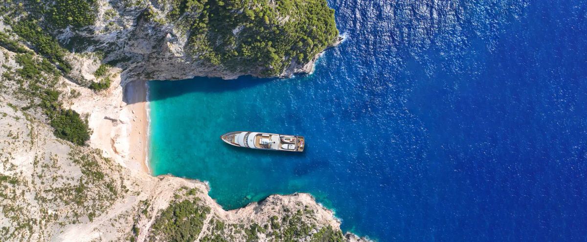 Top-down aerial view of a luxury yacht in a turquoise bay surrounded by cliffs.