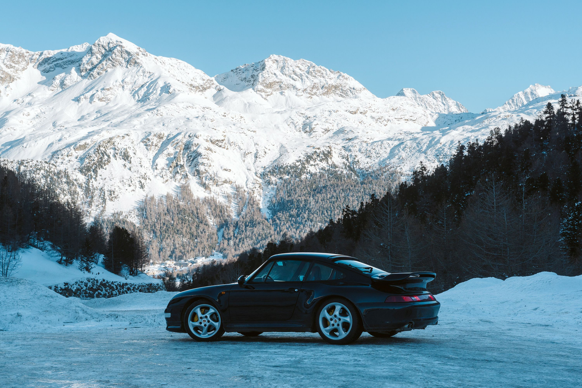 Dark Porsche in side profile on an icy clearing, with forested slopes and sunlit snow-covered mountains behind it., by David Szabo