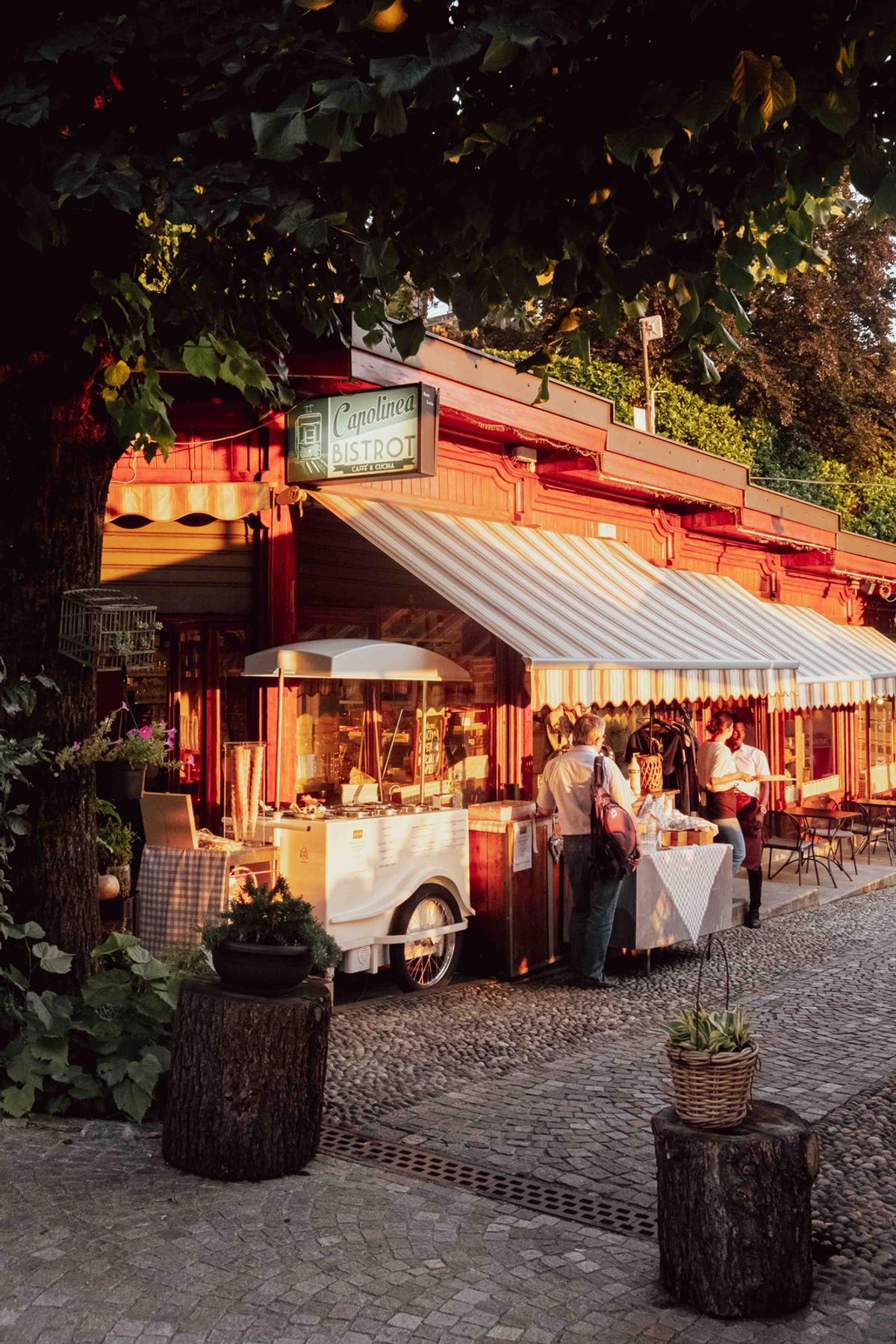 A striped awning opens over a warmly lit café beneath the trees near Lake Como, with red timber and pale tablecloths.
