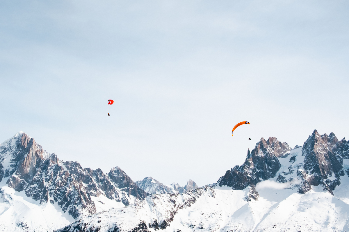 Two paragliders, one red and one orange, fly above a jagged snow-covered mountain range under a wide blue sky.