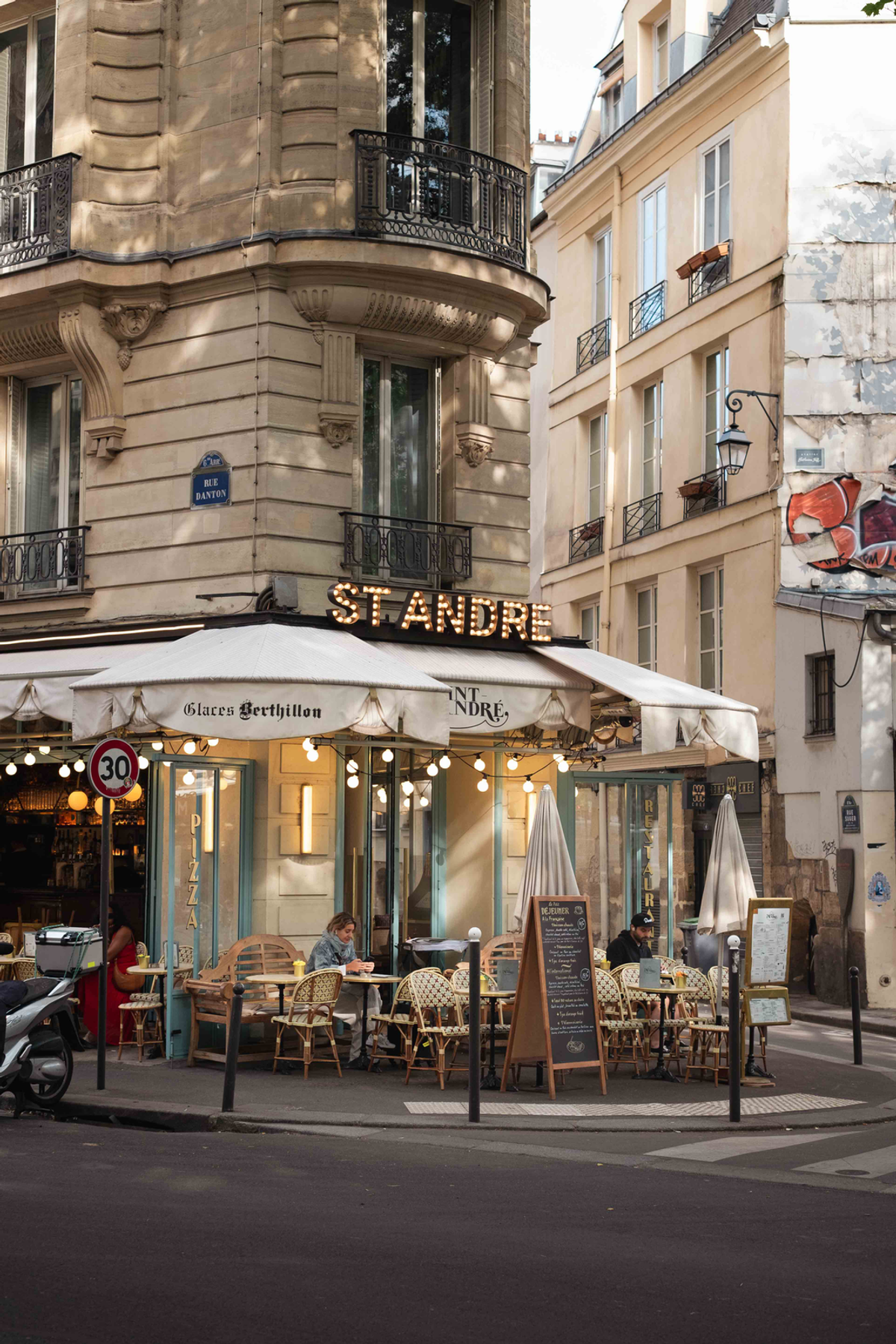 The illuminated Saint André café occupies a curved Paris street corner beneath pale stone apartments, with wicker chairs and diners at pavement tables.