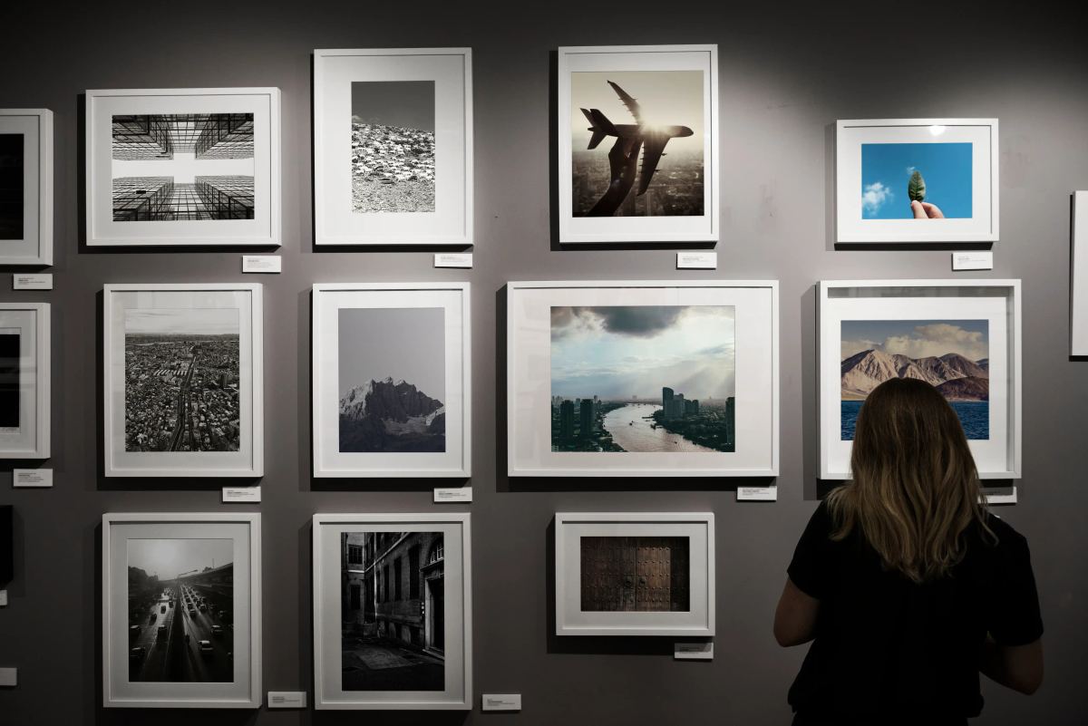 A woman views a gallery wall featuring a grid of framed photographs of landscapes and cityscapes.