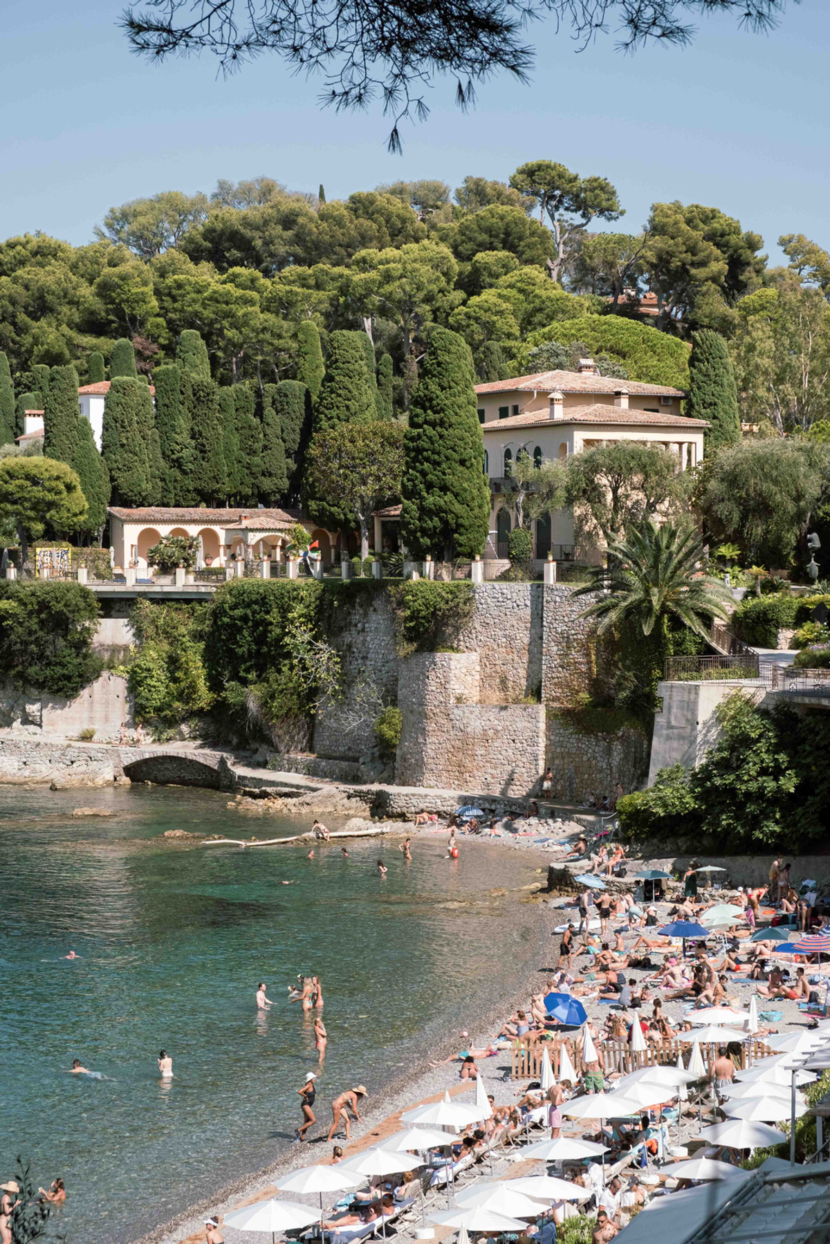 Bathers and white parasols fill a small curved beach below a pale villa, tall clipped cypress trees and a stone seawall beside green-blue water.