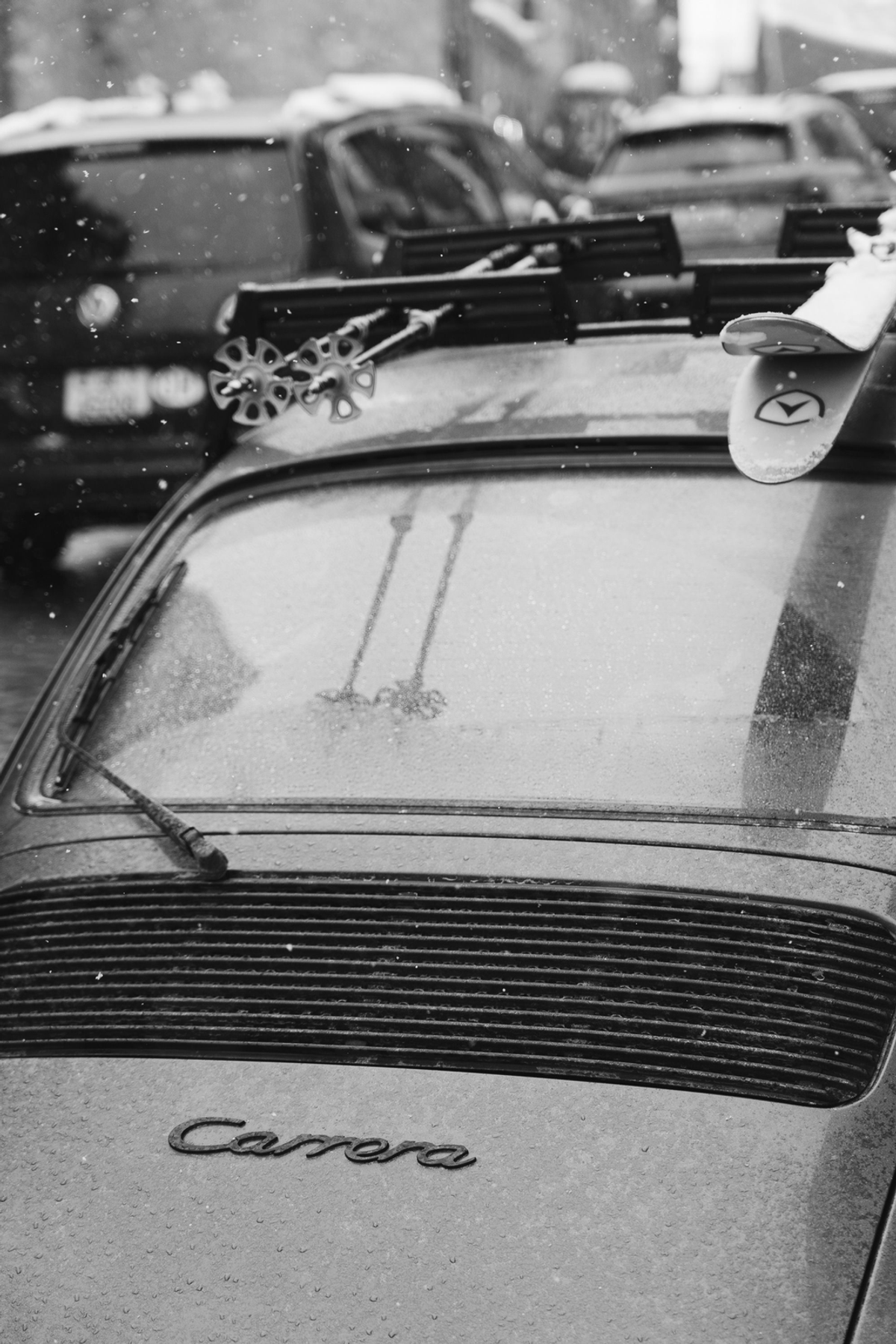 Black-and-white close-up of a snow-dusted Porsche Carrera's rear window and badge, with skis and poles on its roof rack.