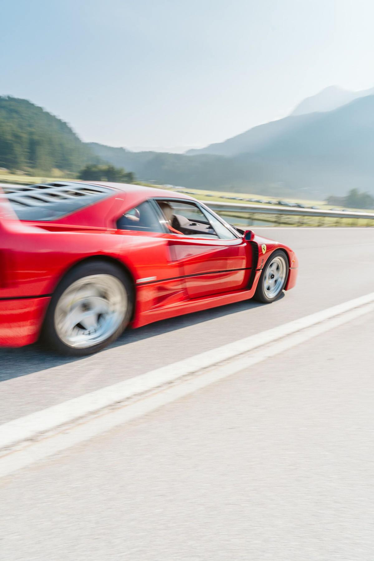 Red Ferrari F40 passing on a mountain road, with blurred wheels, green slopes and a pale blue sky.